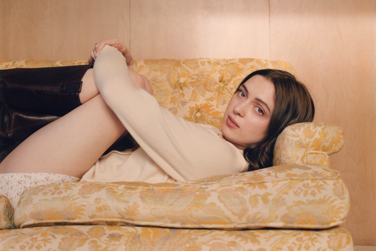 Singer-songwriter Lizzy McAlpine reclines on a vintage floral armchair wearing a white long-sleeve top and boots.